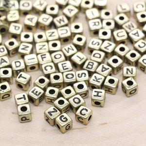 May include: A collection of small, square, gold-colored letter beads scattered on a light wooden surface. Each bead has a single letter printed in black, with various letters visible, ideal for jewelry making or crafting.