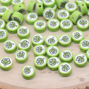 May include: A collection of green and white round beads with a cartoon frog face design. The beads are cylindrical with a hole through the center for stringing. The beads are scattered on a wooden surface.