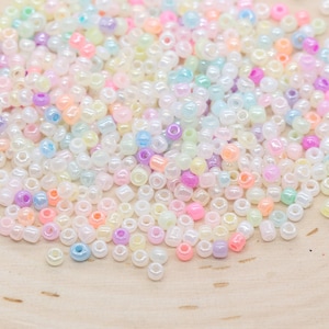 May include: A large assortment of iridescent seed beads in pastel colors, including pink, blue, yellow, and white. The beads are scattered on a wooden surface.