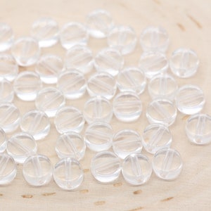 May include: A collection of clear round beads with a hole in the center. The beads are arranged in a random pattern on a light wood surface.