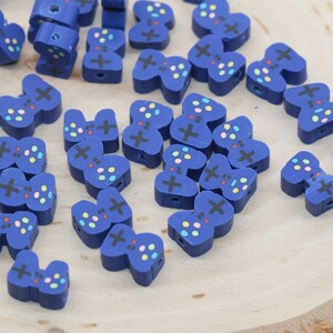 May include: A collection of small, blue wooden beads shaped like video game controllers. Each bead features a painted cross and colorful buttons, with a hole for stringing. The beads are scattered on a light-colored wooden surface.