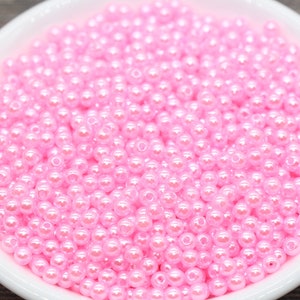 May include: A white, shallow bowl filled with numerous small, round, pink beads. The beads have a glossy, pearl-like finish, reflecting light. The bowl is set against a blurred, neutral-toned background, highlighting the beads' color and texture.