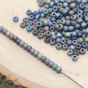 May include: A strand of iridescent blue seed beads strung on a silver wire. The beads are scattered on a wooden surface.