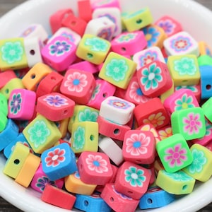 May include: A bowl filled with colourful square-shaped beads with floral designs. The beads are made of polymer clay and have a hole in the centre for stringing.