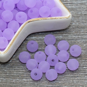 May include: A close-up of a bowl filled with lavender-colored beads. The beads are round and have a frosted finish. Some beads are scattered on a wooden surface.