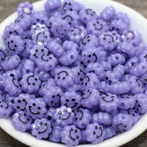 May include: A bowl filled with purple plastic beads. Each bead is shaped like a flower with a smiley face on it.