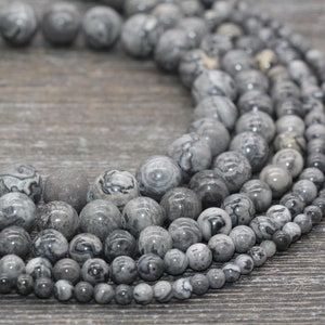 May include: A close-up of a strand of gray and black gemstone beads. The beads are round and have a marbled pattern. The beads are arranged in a circle, with the largest beads in the center and the smallest beads on the outside.