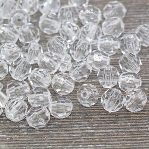 May include: A pile of clear plastic faceted beads. The beads are round and have a hole in the center. They are scattered on a wooden surface.