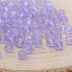 May include: A collection of translucent, round, lavender-colored beads with iridescent shimmer. The beads are scattered on a light-colored wooden surface. Each bead has a hole for stringing.