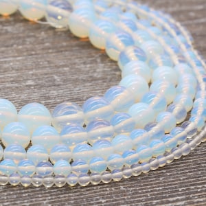May include: A collection of white opal beads in various sizes, arranged in a circular pattern. The beads have a translucent, iridescent quality, with hints of blue and pink.
