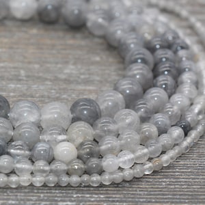 May include: A strand of gray and clear quartz beads. The beads are round and smooth, and they are arranged in a circular pattern. The beads are of varying sizes, with the larger beads in the center and the smaller beads on the outside.