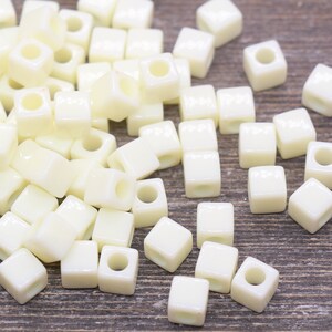 May include: A pile of small, white, square beads with a hole in the center. The beads are made of ceramic and have a glossy finish.