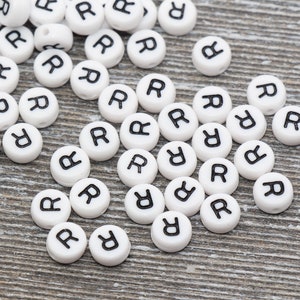 May include: A collection of white, round beads with the letter "R" printed in black. The beads are scattered on a gray wooden surface, ideal for crafting or jewelry making.