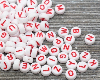 May include: White plastic alphabet beads with red letters. The beads are scattered on a wooden surface.