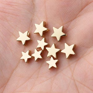 May include: A collection of nine small, gold-colored star-shaped beads with a smooth, metallic finish. Each star features five points and a flat surface, arranged in a cluster. These components are designed for jewelry making and crafting projects.