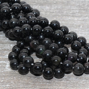 May include: A strand of black obsidian beads, each bead is round and smooth. The beads are arranged in a loose pile on a wooden surface.