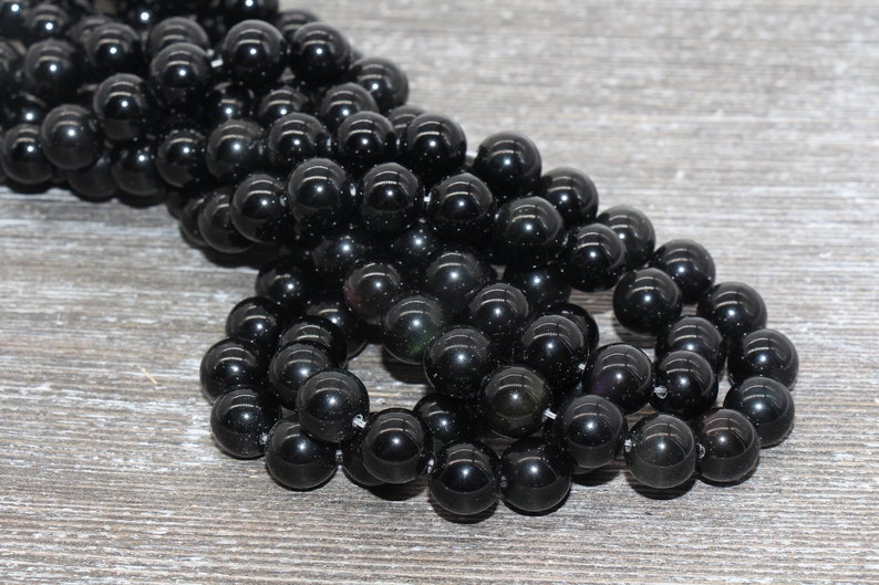 May include: A strand of black obsidian beads, each bead is round and smooth. The beads are arranged in a loose pile on a wooden surface.