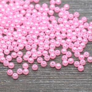 May include: A collection of small, pink, round beads scattered on a wooden surface. The beads have a glossy finish and are evenly sized. These beads are suitable for jewelry making, crafts, and other creative projects.