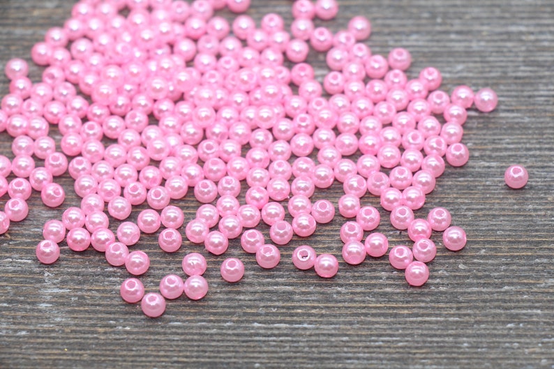May include: A collection of small, pink, round beads scattered on a wooden surface. The beads have a glossy finish and are evenly sized. These beads are suitable for jewelry making, crafts, and other creative projects.