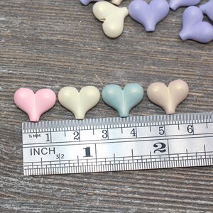 May include: Five pastel-colored heart-shaped buttons, measuring approximately 1 inch in diameter, are lined up on a ruler. The buttons are pink, cream, light blue, beige, and lavender.