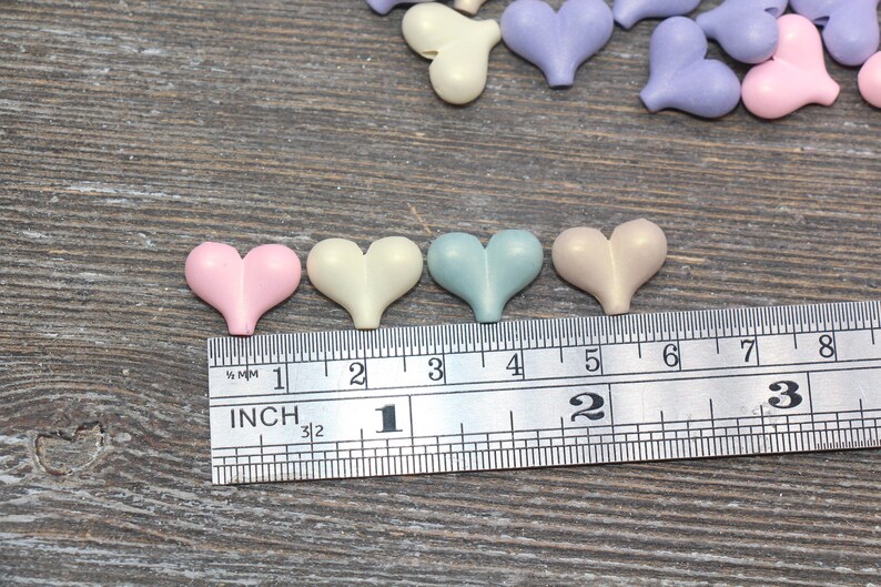 May include: Five pastel-colored heart-shaped buttons, measuring approximately 1 inch in diameter, are lined up on a ruler. The buttons are pink, cream, light blue, beige, and lavender.