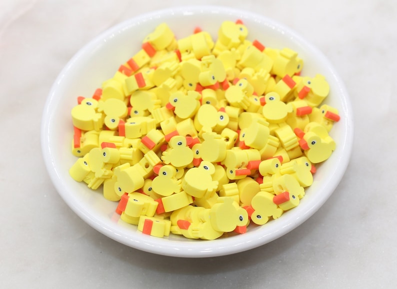 May include: A bowl filled with yellow rubber ducky shaped charms with orange beaks. The charms are perfect for adding a playful touch to crafts and jewellery.