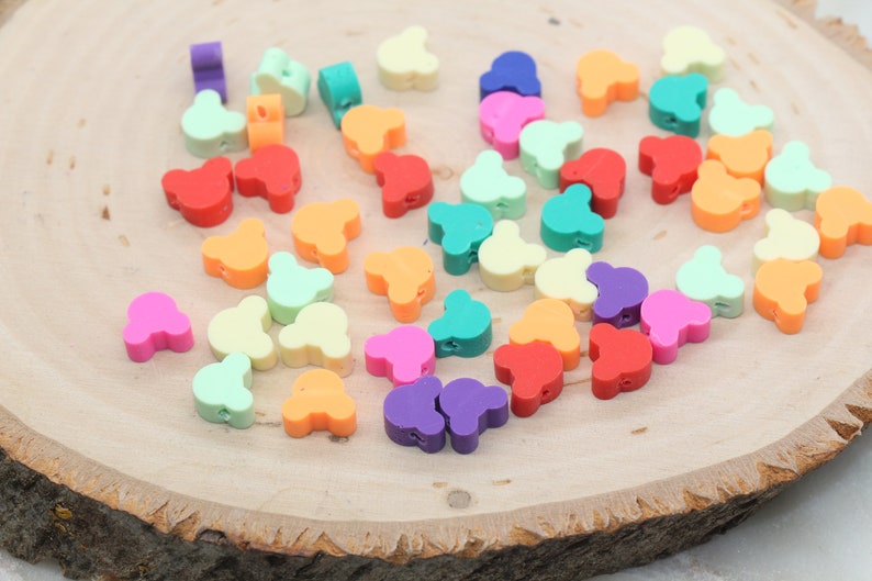 May include: Assortment of colourful plastic bear-shaped beads in various colours, including red, orange, yellow, green, blue, purple, and pink. The beads are arranged on a wooden surface.
