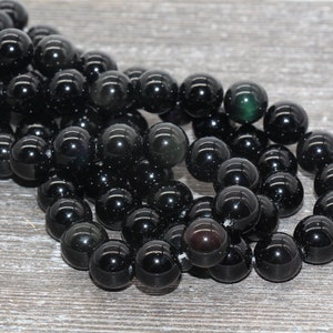 May include: A strand of black obsidian beads with a slight green shimmer. The beads are round and smooth, and they are arranged in a loose pile.