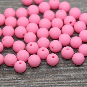 May include: A large group of pink silicone beads, scattered on a wooden surface. The beads are round and smooth, with a small hole in the center of each bead.
