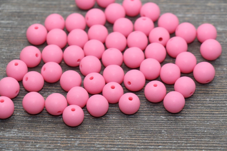 May include: A large group of pink silicone beads, scattered on a wooden surface. The beads are round and smooth, with a small hole in the center of each bead.