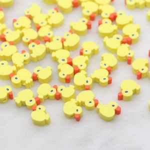 May include: A large group of yellow rubber ducky charms with orange beaks. The ducky charms are scattered on a white surface.