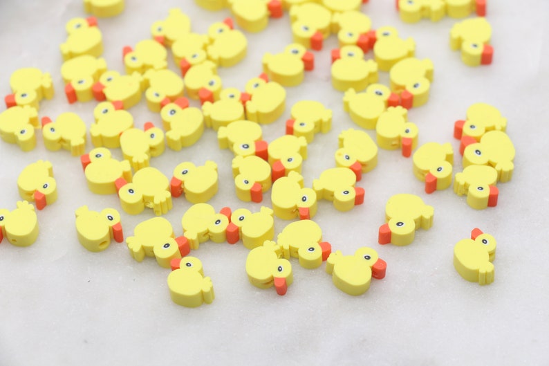 May include: A large group of yellow rubber ducky charms with orange beaks. The ducky charms are scattered on a white surface.
