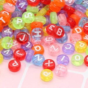 May include: A large assortment of colourful alphabet letter beads. The beads are made of clear plastic with white letters. The beads are scattered on a pink surface.