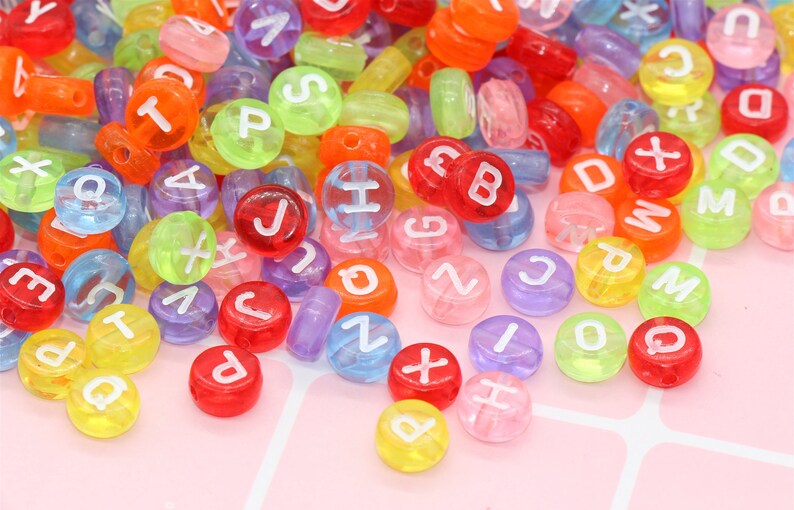 May include: A large assortment of colourful alphabet letter beads. The beads are made of clear plastic with white letters. The beads are scattered on a pink surface.