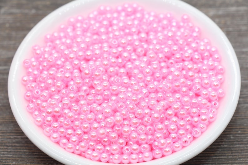 May include: A white, shallow bowl filled with numerous small, round, pink beads. The beads have a glossy, pearl-like finish, reflecting light. The bowl is set against a blurred, neutral-toned background, highlighting the beads' color and texture.