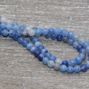 Pode incluir: Close-up de contas de pedras preciosas azuis em tons variados de azul e branco. As contas s&atilde;o redondas e lisas, com um acabamento brilhante. As contas s&atilde;o enfiadas, criando um colar ou uma pulseira.