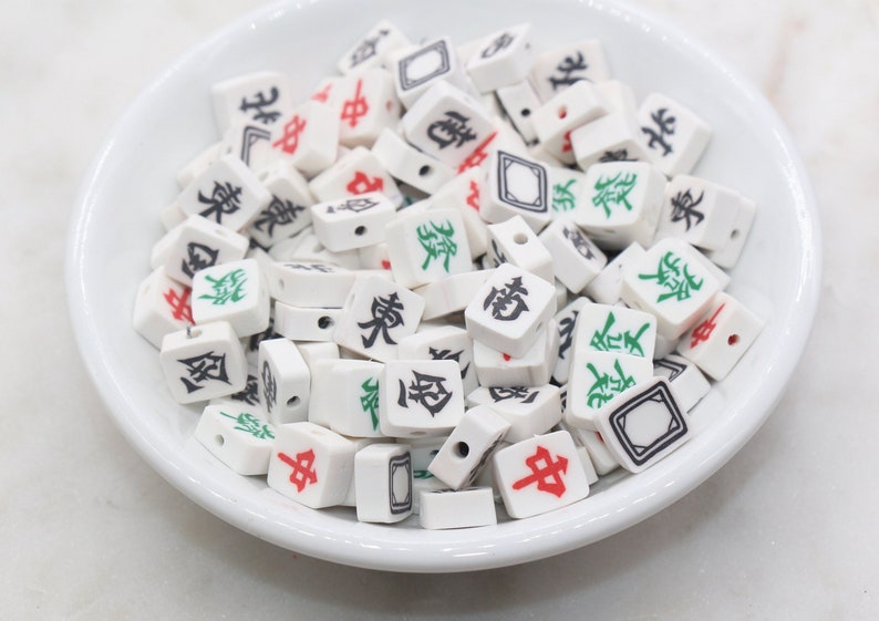 K&ouml;nnte beinhalten: Eine wei&szlig;e Sch&uuml;ssel gef&uuml;llt mit wei&szlig;en Keramik-Mahjong-Steinen. Die Steine haben schwarze, rote und gr&uuml;ne chinesische Schriftzeichen und Symbole.