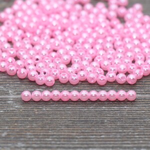 May include: A collection of small, round, light pink beads scattered on a wooden surface. A single strand of the same beads is arranged in a straight line. The beads have a glossy finish, reflecting light.