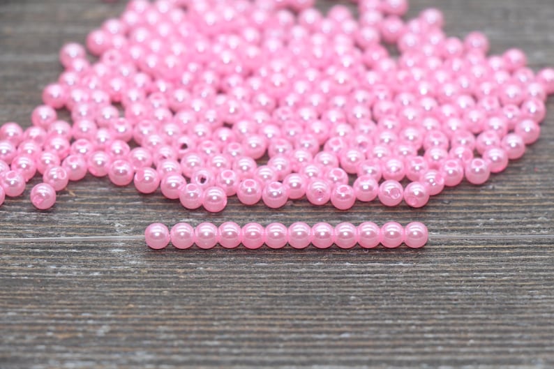May include: A collection of small, round, light pink beads scattered on a wooden surface. A single strand of the same beads is arranged in a straight line. The beads have a glossy finish, reflecting light.