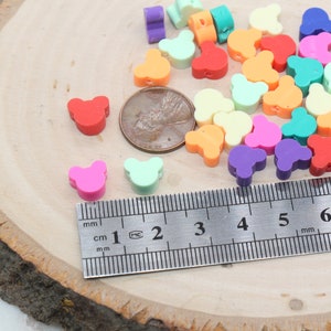 May include: Assortment of colourful plastic bear-shaped beads, measuring approximately 1 cm in diameter, displayed on a wooden surface with a ruler for scale.