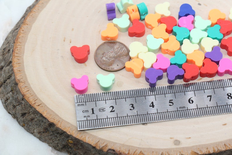 May include: Assortment of colourful plastic bear-shaped beads, measuring approximately 1 cm in diameter, displayed on a wooden surface with a ruler for scale.