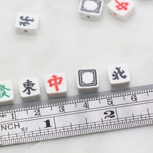K&ouml;nnte beinhalten: Wei&szlig;e chinesische Mahjong-Steine mit schwarzen und roten Schriftzeichen auf wei&szlig;em Hintergrund. Die Steine sind auf einem Lineal mit Zollmarkierungen ausgerichtet. Die Steine zeigen "發", "東", "中".