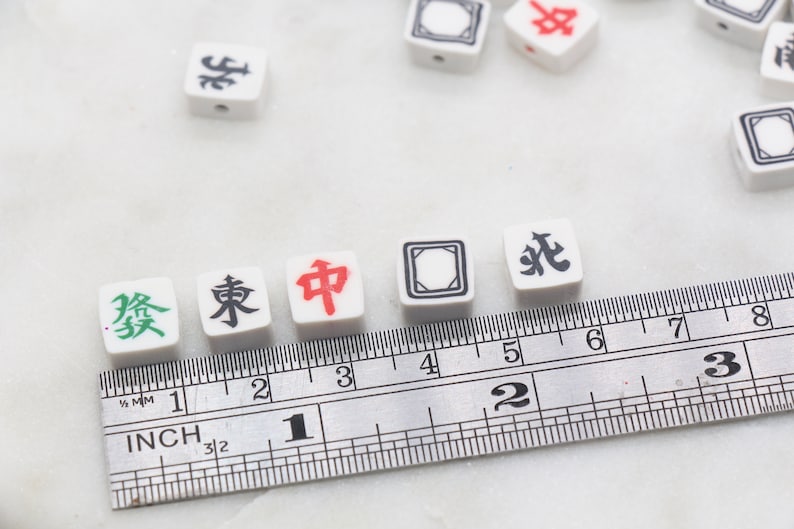 K&ouml;nnte beinhalten: Wei&szlig;e chinesische Mahjong-Steine mit schwarzen und roten Schriftzeichen auf wei&szlig;em Hintergrund. Die Steine sind auf einem Lineal mit Zollmarkierungen ausgerichtet. Die Steine zeigen "發", "東", "中".