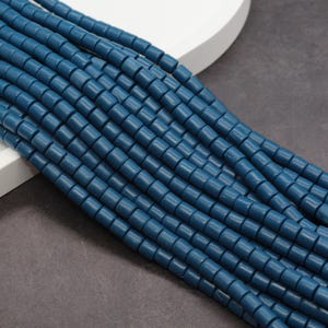 May include: Strands of teal blue, square-shaped beads. The beads are strung together, creating a textured, linear pattern. The beads are arranged diagonally across the frame, with a white surface and a dark gray surface in the background.
