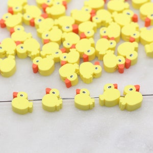 May include: A group of yellow rubber ducky charms with orange beaks. The charms are scattered on a white surface. A few of the charms are strung on a silver wire.