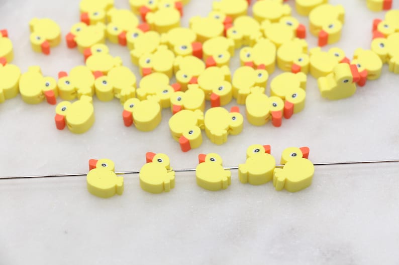 May include: A group of yellow rubber ducky charms with orange beaks. The charms are scattered on a white surface. A few of the charms are strung on a silver wire.