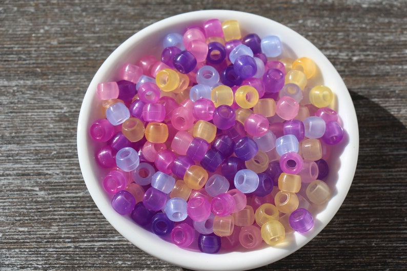 Pode incluir: Uma tigela branca cheia de contas de pl&aacute;stico transl&uacute;cidas coloridas. As contas s&atilde;o em tons de amarelo, rosa, roxo e azul.