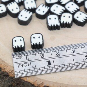 May include: Black and white ghost-shaped beads with a ruler for size reference. The beads are approximately 2.5 cm in size.