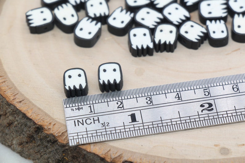 May include: Black and white ghost-shaped beads with a ruler for size reference. The beads are approximately 2.5 cm in size.