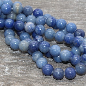 Pode incluir: Fio de contas redondas de pedra azul. As contas t&ecirc;m uma superf&iacute;cie lisa e polida e variam em tons de azul, do claro ao &iacute;ndigo profundo. Est&atilde;o enfiadas, adequadas para bijuteria. O fundo &eacute; uma superf&iacute;cie de madeira cinza envelhecida.